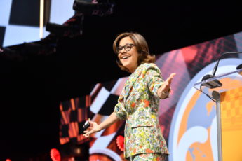 Woman with brown hair and glasses in a floral suit on stage.