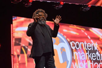 Black man with a mustache and beard wearing a black shirt, sport coat, and black pants on stage.